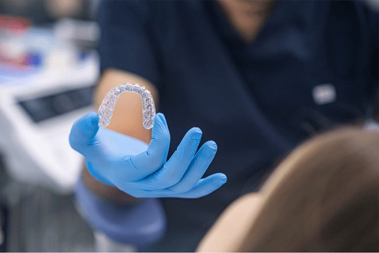A dentist in blue gloves holds a clear dental aligner over a patient reclined in a dental chair.