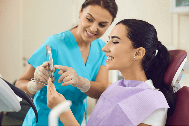 A woman sits in a dental chair while another woman in a blue shirt stands beside her, likely providing assistance.