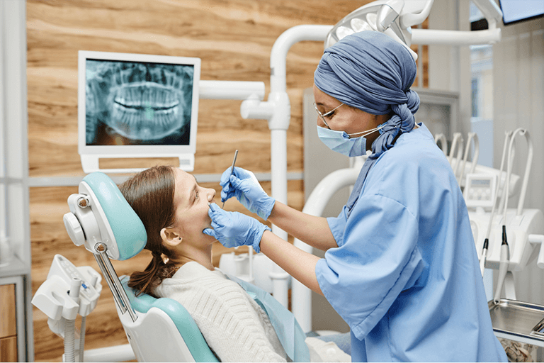 A dentist is cleaning a woman's teeth in a dental office, with tools and equipment visible in the background.